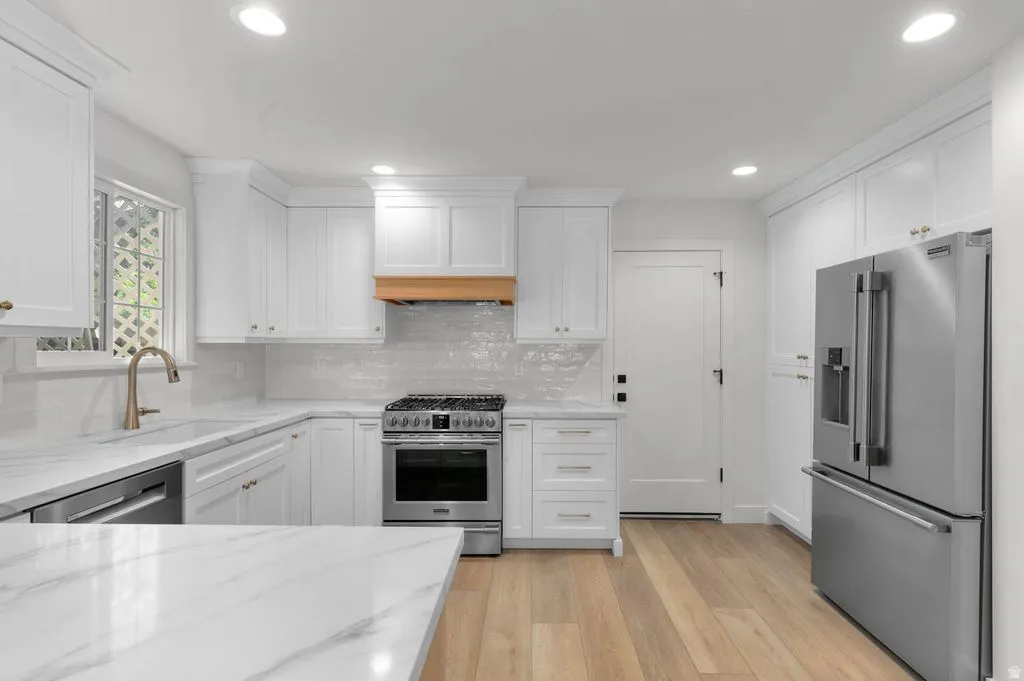White kitchen featuring a gas range, stainless steel appliances, marble-look countertops, white cabinetry with gold-toned hardware, and light wood-finish flooring