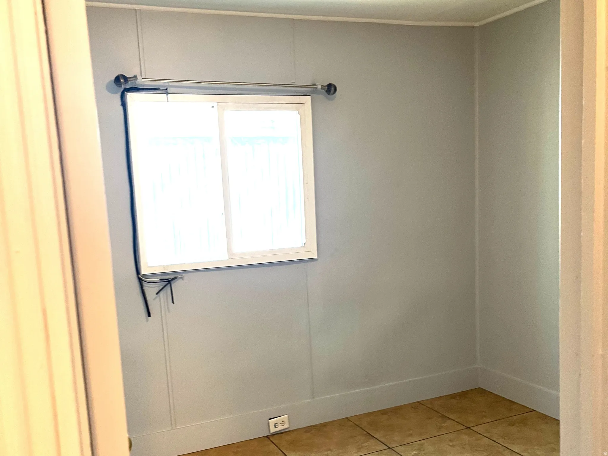 Room featuring neutral-toned tile flooring, light gray wall paneling, and a single window with a dark curtain rod