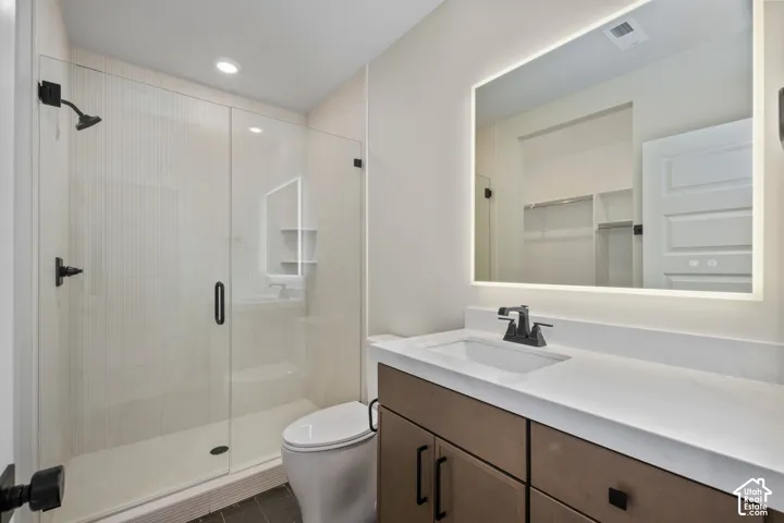 Contemporary bathroom featuring a frameless glass shower enclosure, a single vanity with a white countertop and wood-finish cabinetry, a lighted mirror, and matte black fixtures