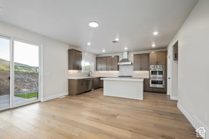 Open-concept living area featuring wood-finish flooring, a contemporary kitchen with dark wood cabinetry, a white island, stainless steel double ovens, and a stainless steel range hood