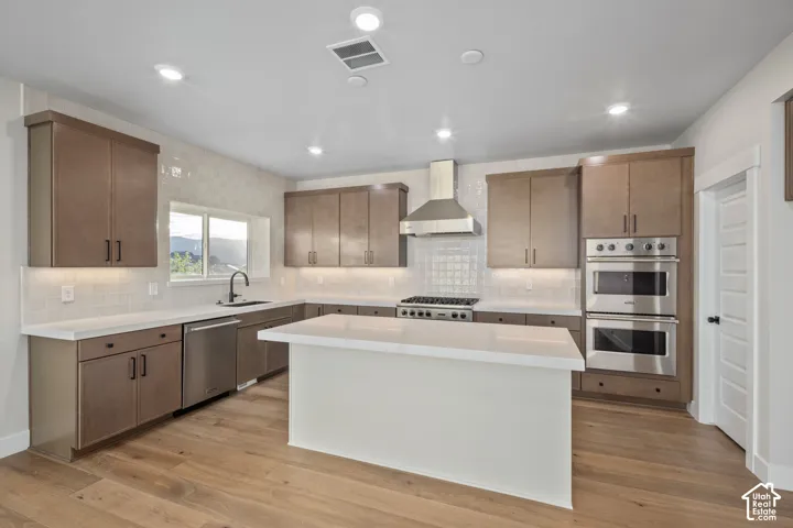 Modern kitchen featuring light wood-finish flooring, a central island with a solid surface countertop, stainless steel appliances including a built-in double oven, and recessed lighting throughout