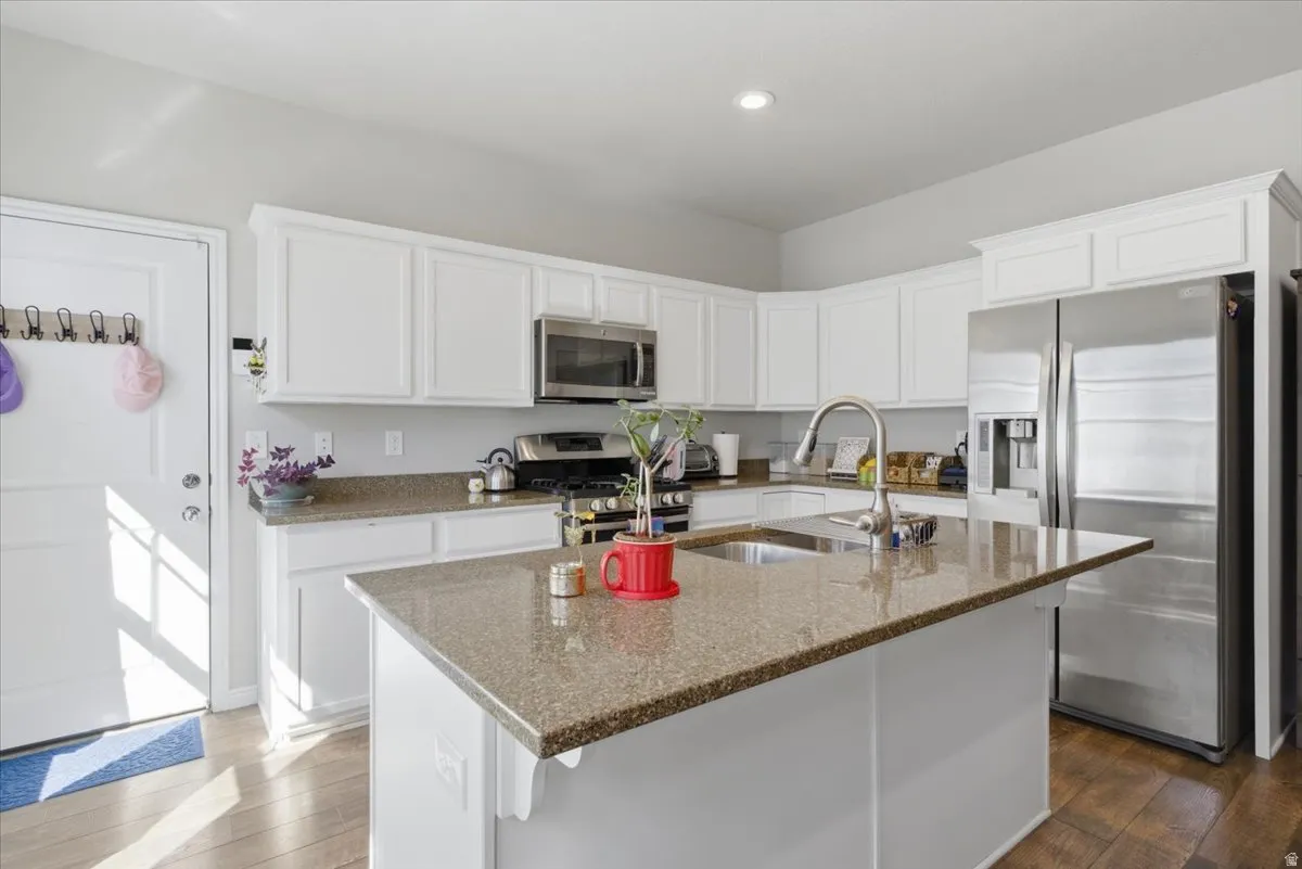 Kitchen featuring a central island with an undermount sink, white cabinetry, integrated stainless steel appliances, speckled countertops, and wood-finish flooring
