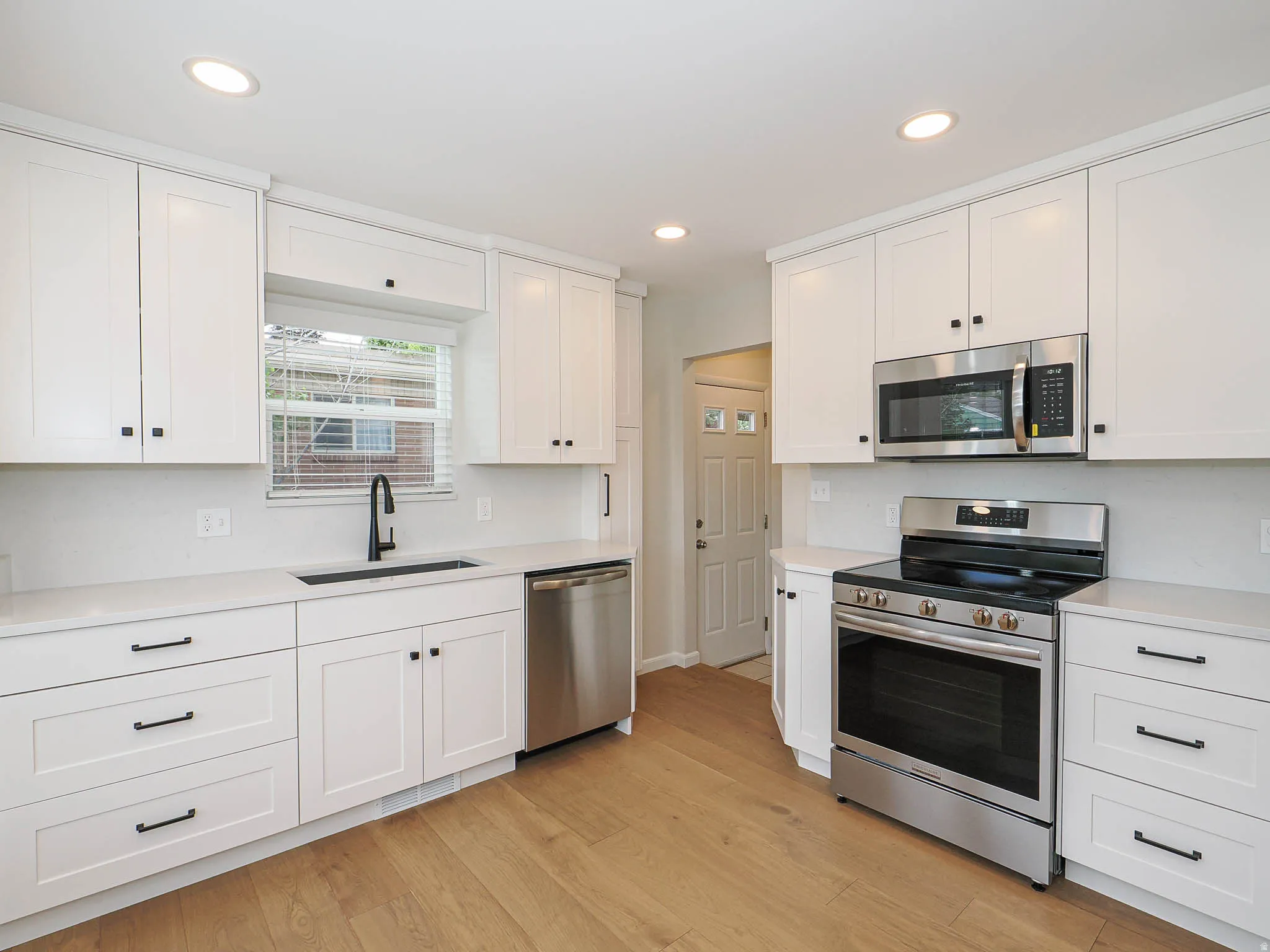 Kitchen featuring white shaker cabinetry, stainless steel appliances, light-toned wood-finish flooring, a matte black kitchen faucet, and a large sink basin