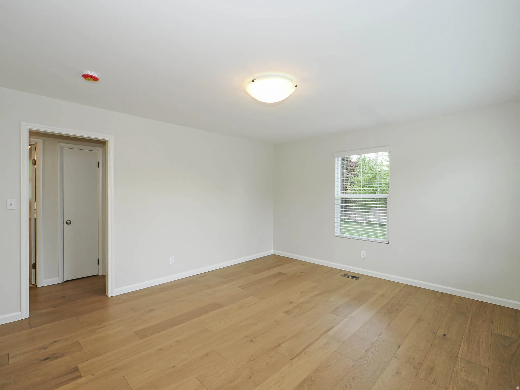 Light wood-finish flooring extending throughout the room, white baseboards, white walls, flush-mount ceiling light fixture, and a window with horizontal blinds