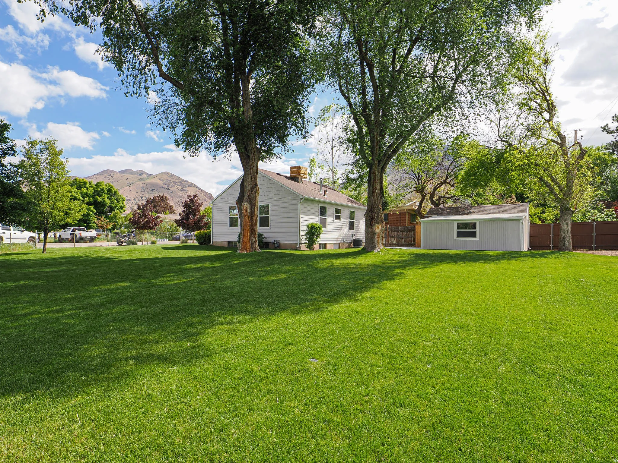 Expansive green lawn with mature trees, a white-sided residence, a detached light gray storage shed with white trim, and a partial mountain backdrop