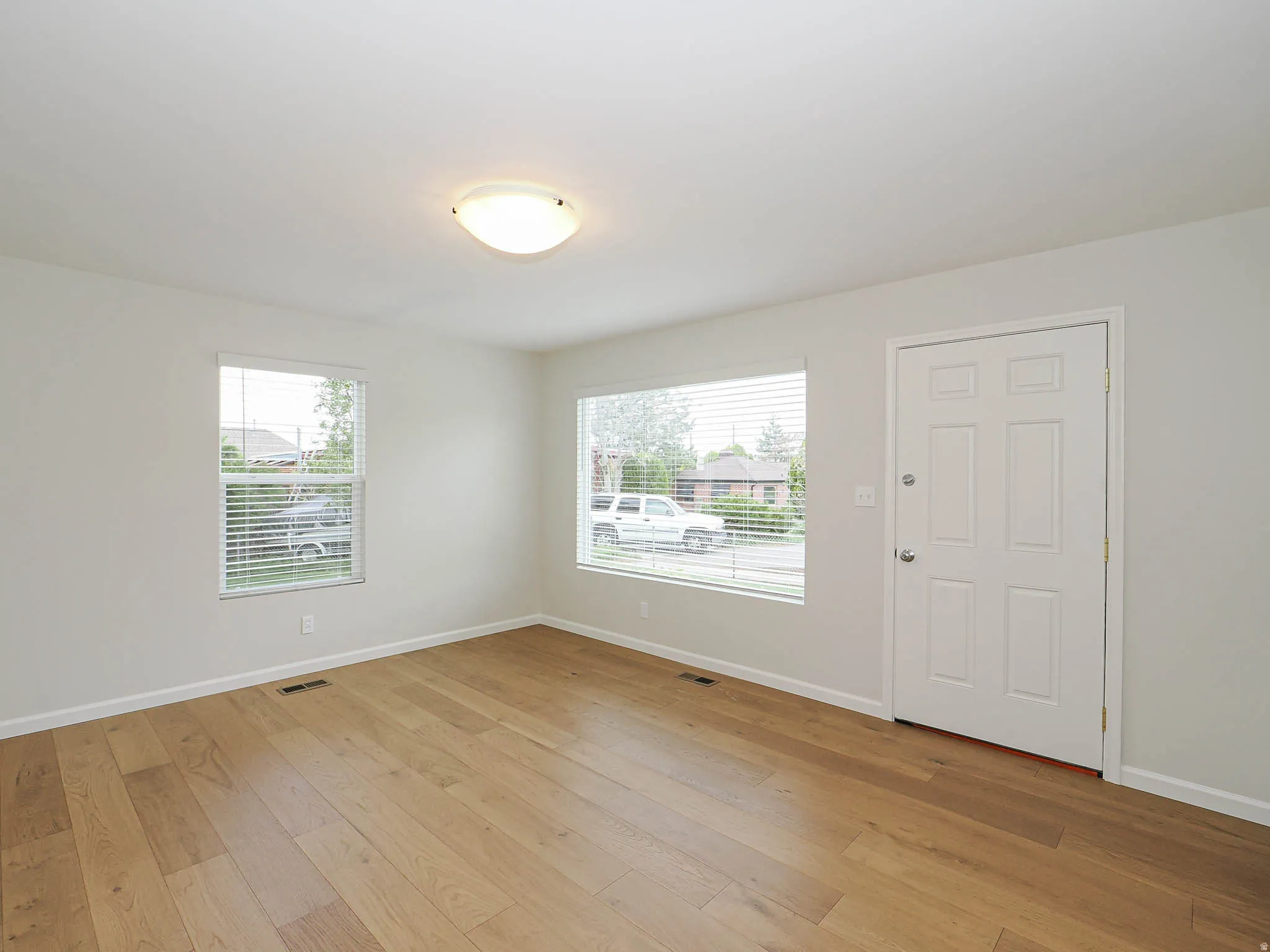 Spacious room featuring wood-finish flooring, light-toned walls, multiple windows with blinds, a flush-mount ceiling light fixture, and a paneled entry door