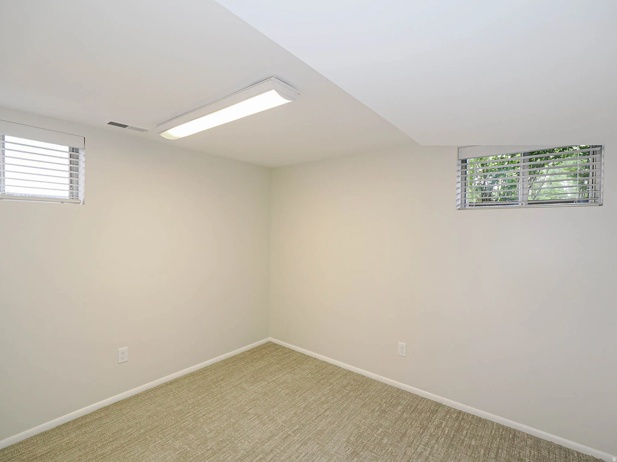 Carpeted room featuring two high-set windows with horizontal blinds, recessed baseboard trim, neutral wall tones, and a ceiling-mounted LED lighting fixture