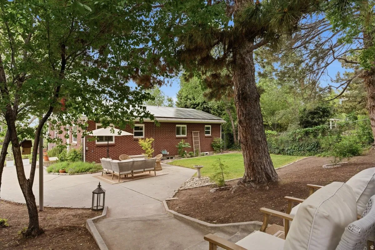 Spacious backyard featuring an extensive concrete patio, mature trees providing natural shade, a manicured lawn area, a brick exterior, and rooftop solar panels