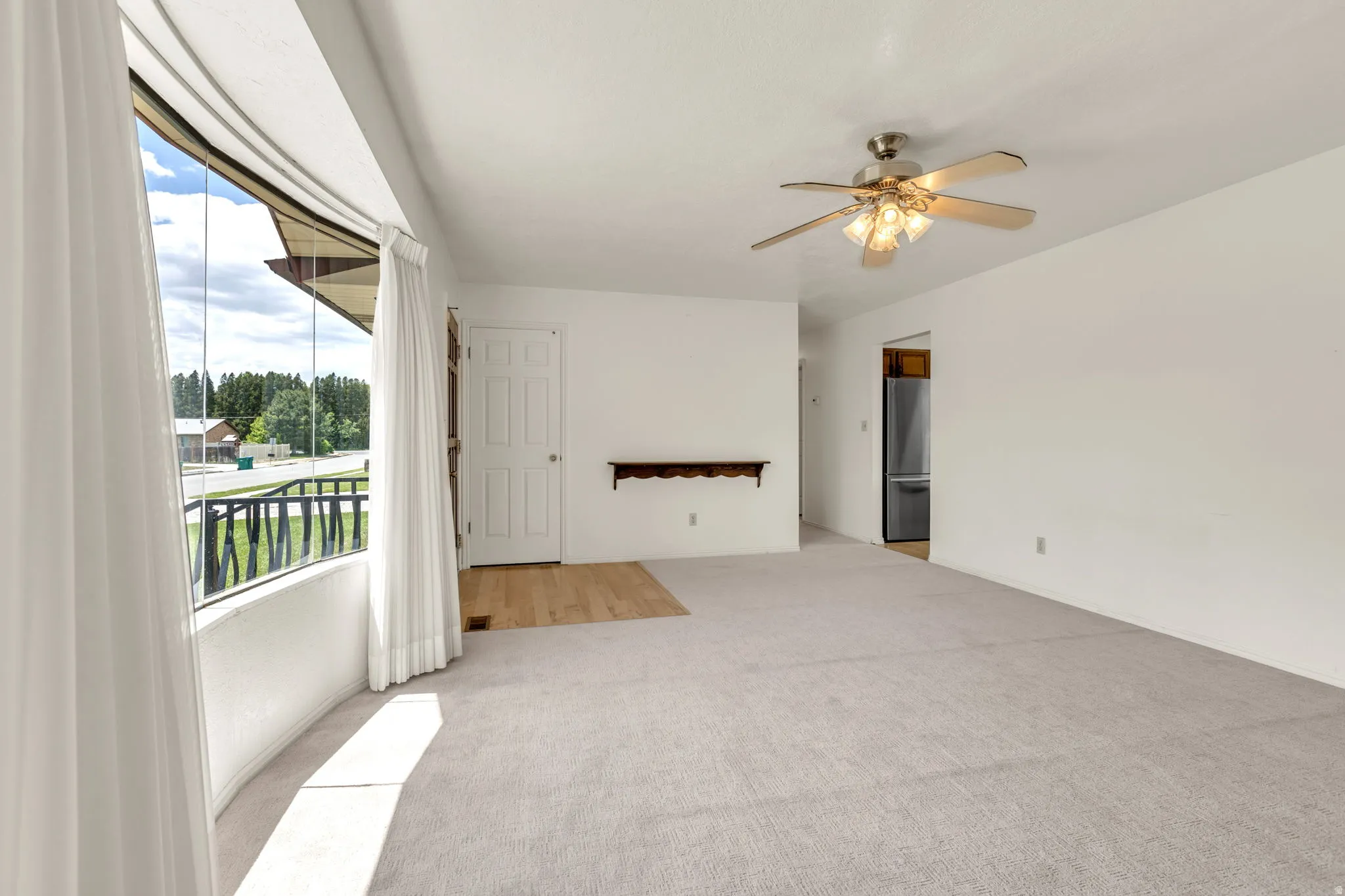 Spacious living area featuring a curved bay window, light-toned carpeting, wood-finish flooring, a ceiling fan with integrated lighting, and a paneled entry door