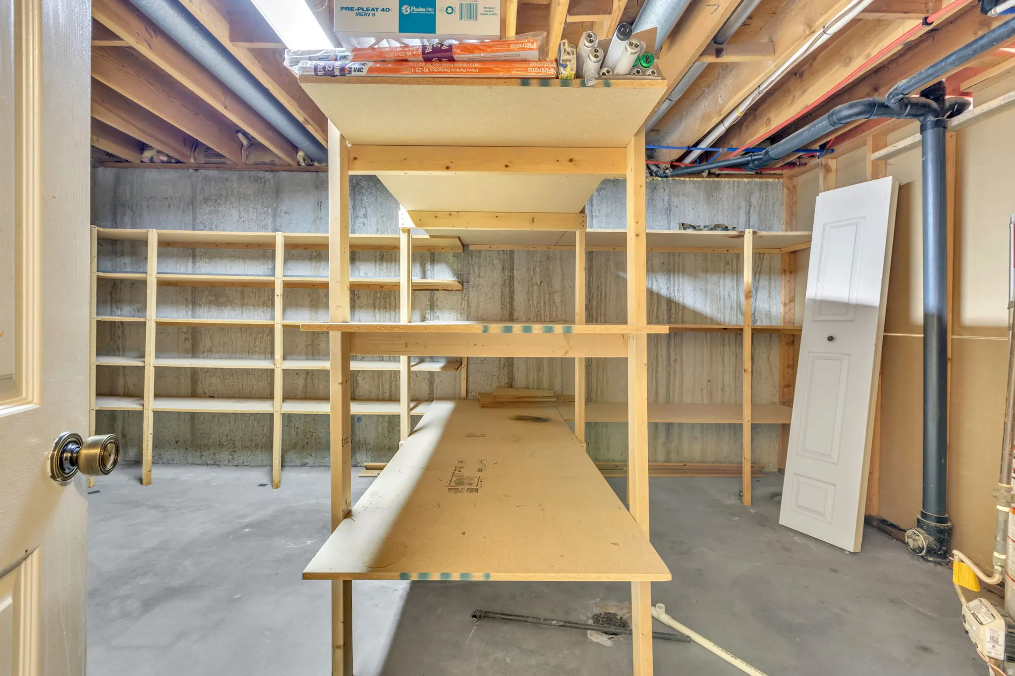 Unfinished basement space featuring exposed ceiling joists, concrete walls, concrete flooring, and extensive built-in wooden shelving for storage