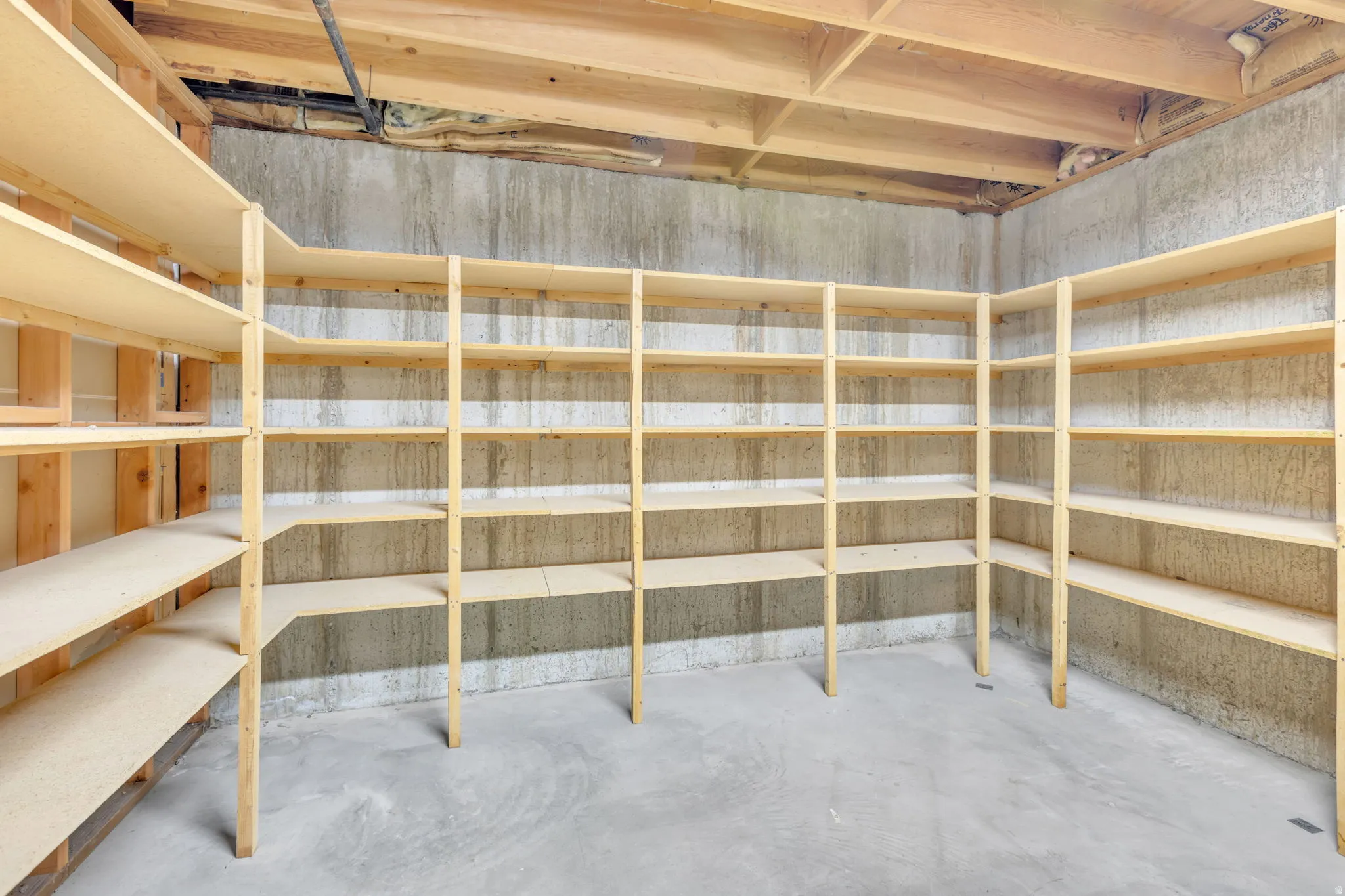 Basement storage area featuring extensive built-in wooden shelving, concrete walls, exposed ceiling joists, and a concrete floor