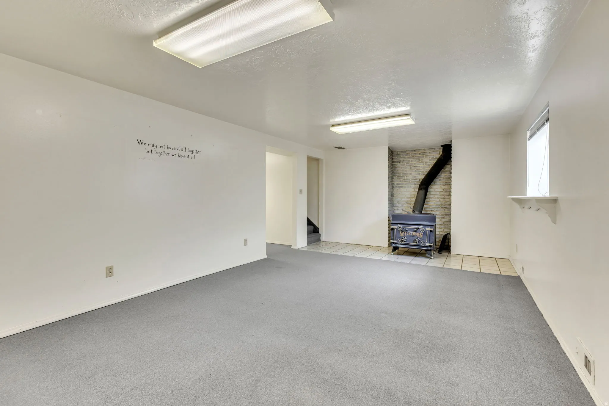 Spacious room featuring gray carpet, a wood-burning stove with a brick surround, and recessed fluorescent lighting