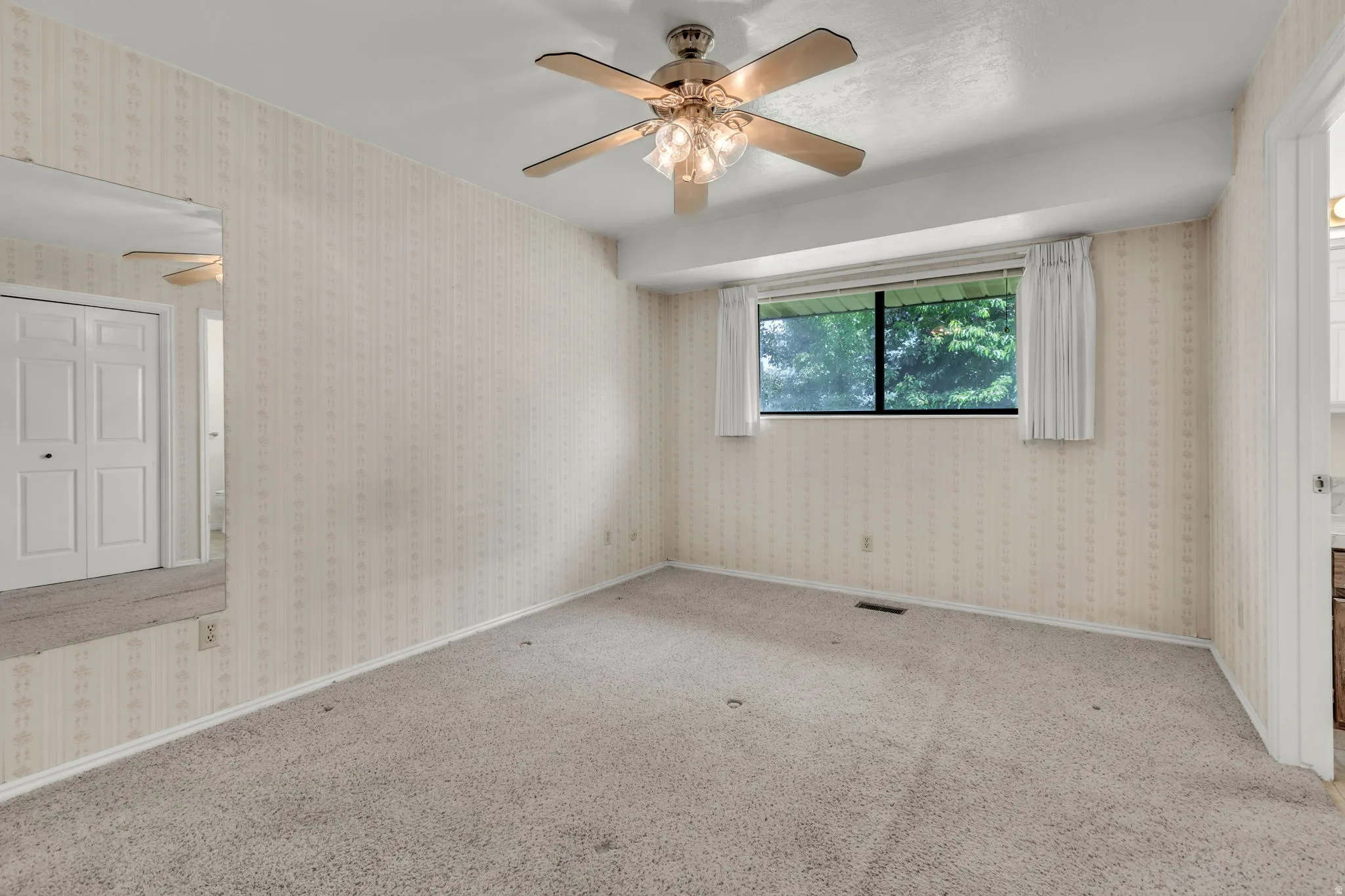 Carpeted room featuring a ceiling fan with integrated lighting, a large window, patterned wallpaper, and a mirrored wall panel
