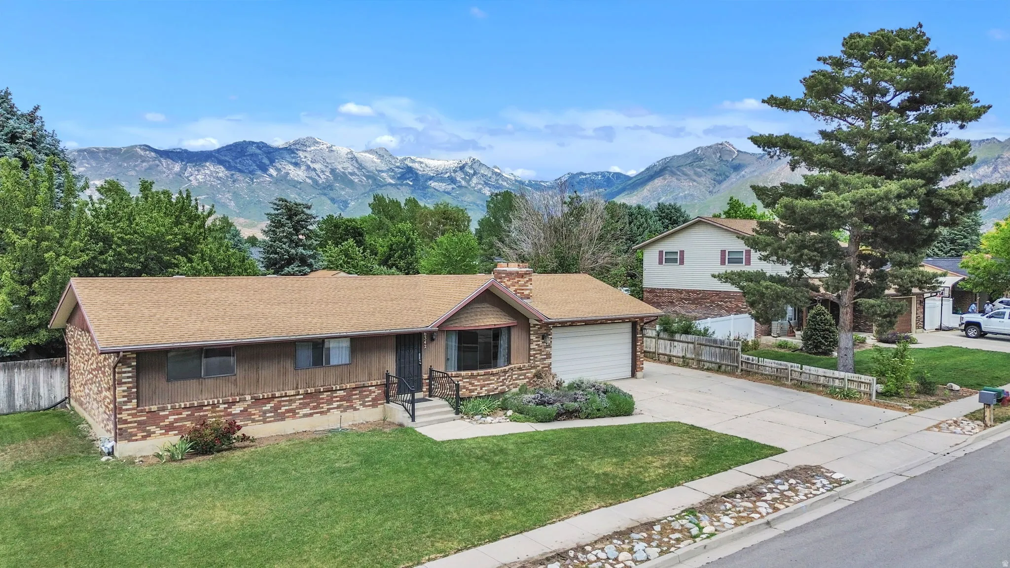 Mountain backdrop property featuring a brick and wood-sided exterior, attached garage, shingle roof, and a mature front yard tree