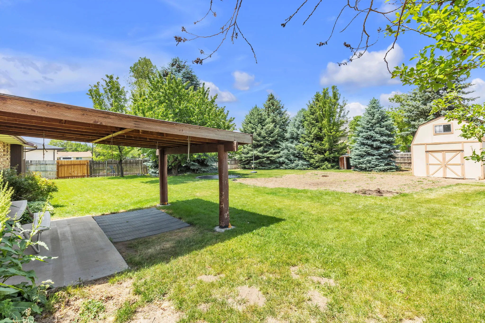 Expansive backyard featuring a covered patio with concrete and paver flooring, a manicured lawn, an auxiliary storage shed with double doors, and mature evergreen trees bordering the property