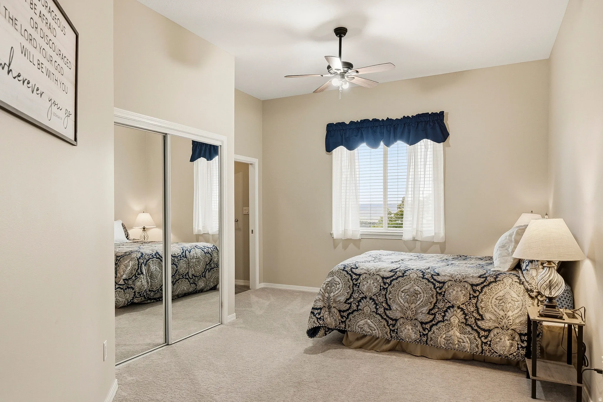 Carpeted interior space featuring a window with integrated blinds, a ceiling fan with light fixture, mirrored closet doors, and neutral wall tones