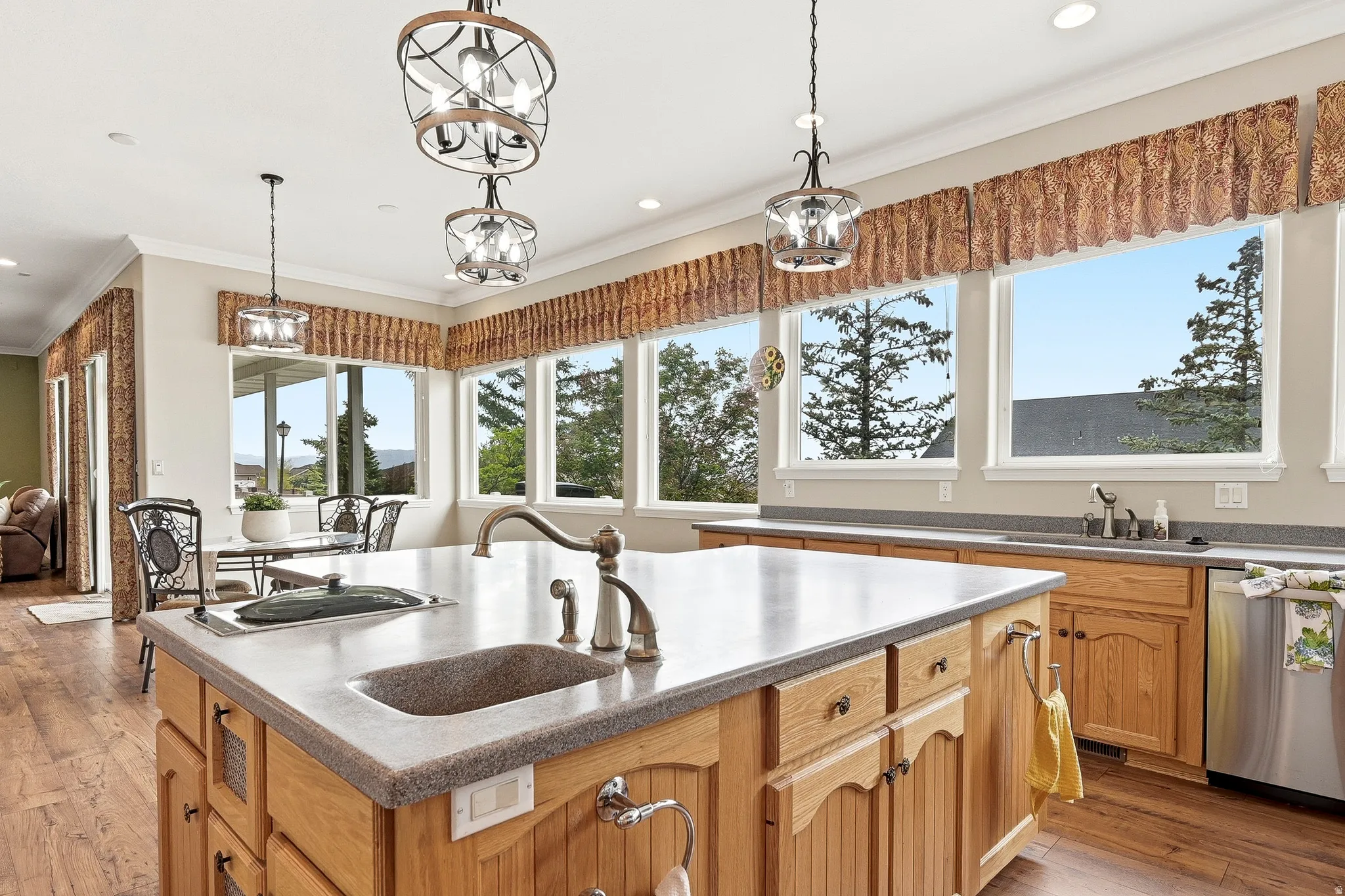 Kitchen featuring a substantial island with an integrated sink and faucet, wood-finish cabinetry, light-toned countertops, expansive windows, and distinctive pendant lighting fixtures