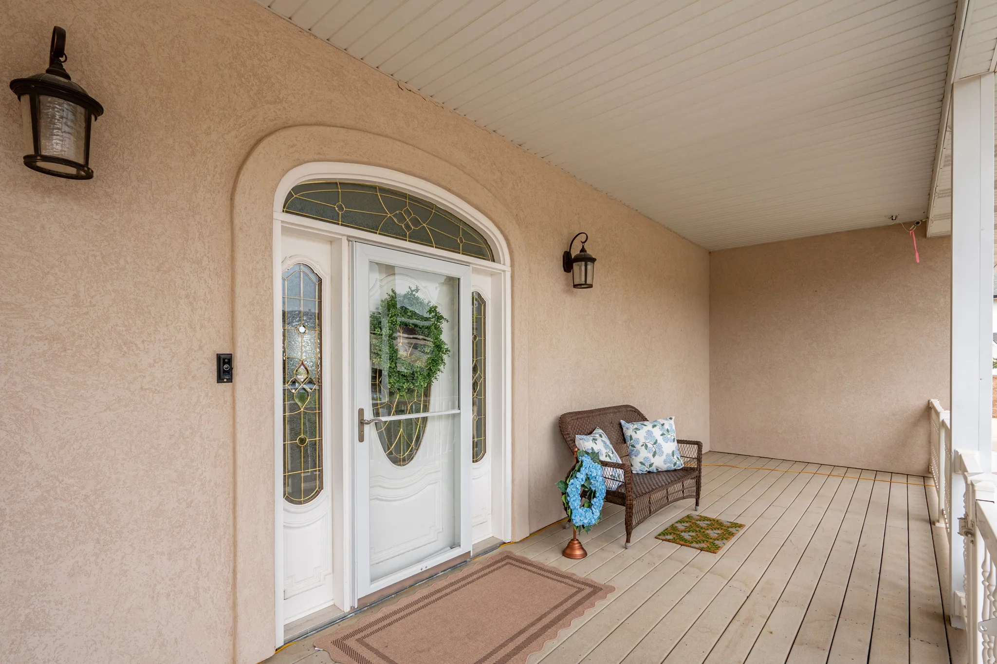 Covered porch entry featuring a white arched door with decorative glass sidelights, a complementary storm door, textured stucco siding, and ceiling-mounted soffit panels