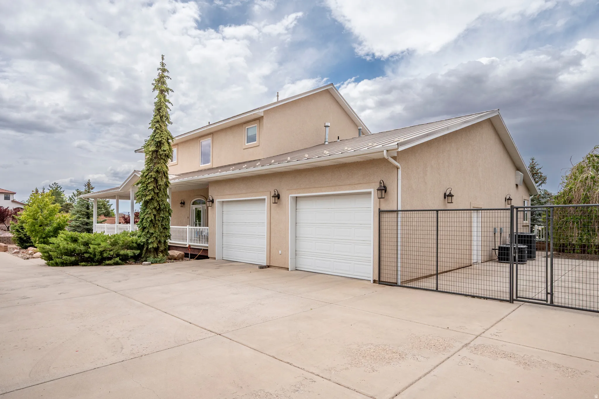 Stucco exterior residence featuring a wide concrete driveway, an attached two-car garage with overhead doors, a covered porch with white railings, and a fenced side yard enclosure