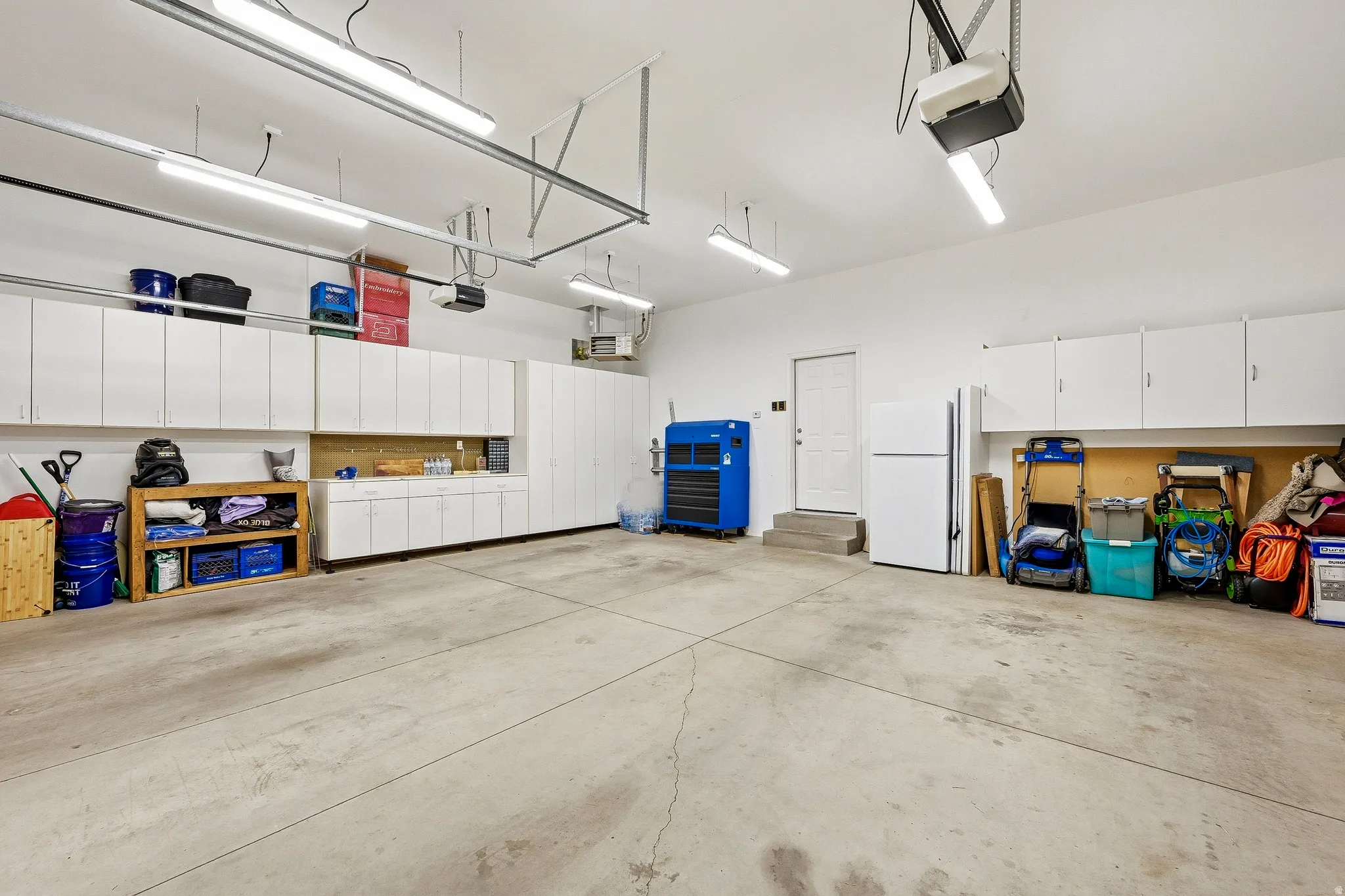 Concrete flooring garage featuring extensive white cabinetry, a built-in workbench area, multiple overhead fluorescent light fixtures, and an automatic garage door opener