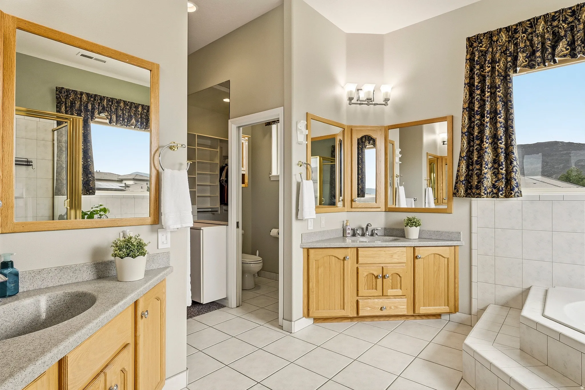 Spacious bathroom featuring dual vanities with light wood cabinetry and light-toned countertops, a glass-enclosed shower, a built-in bathtub, and white tile flooring