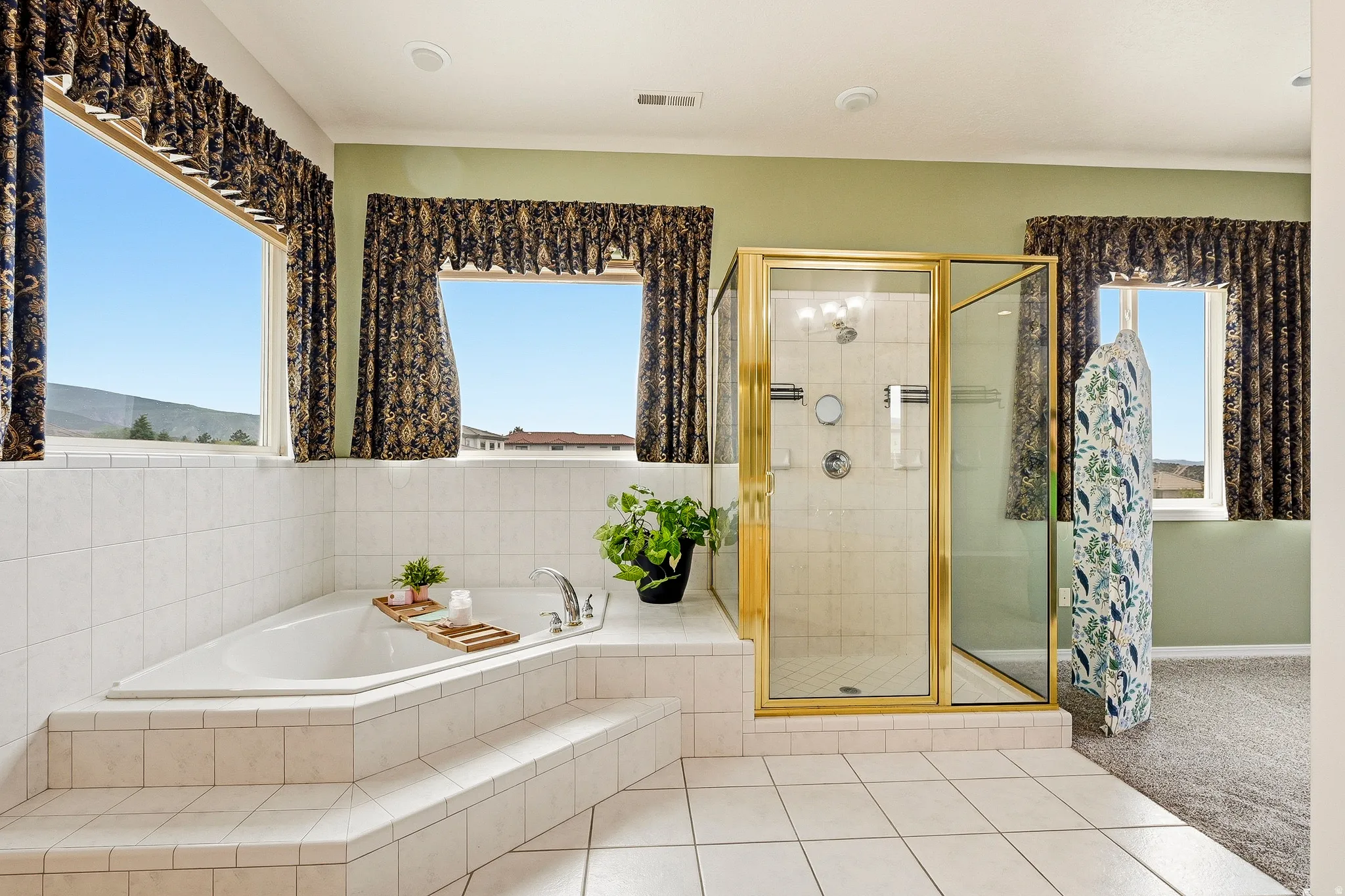 Spacious bathroom featuring a built-in corner soaking tub with tiled surround and steps, a glass-enclosed shower with gold-tone framing, white tile flooring, and two windows
