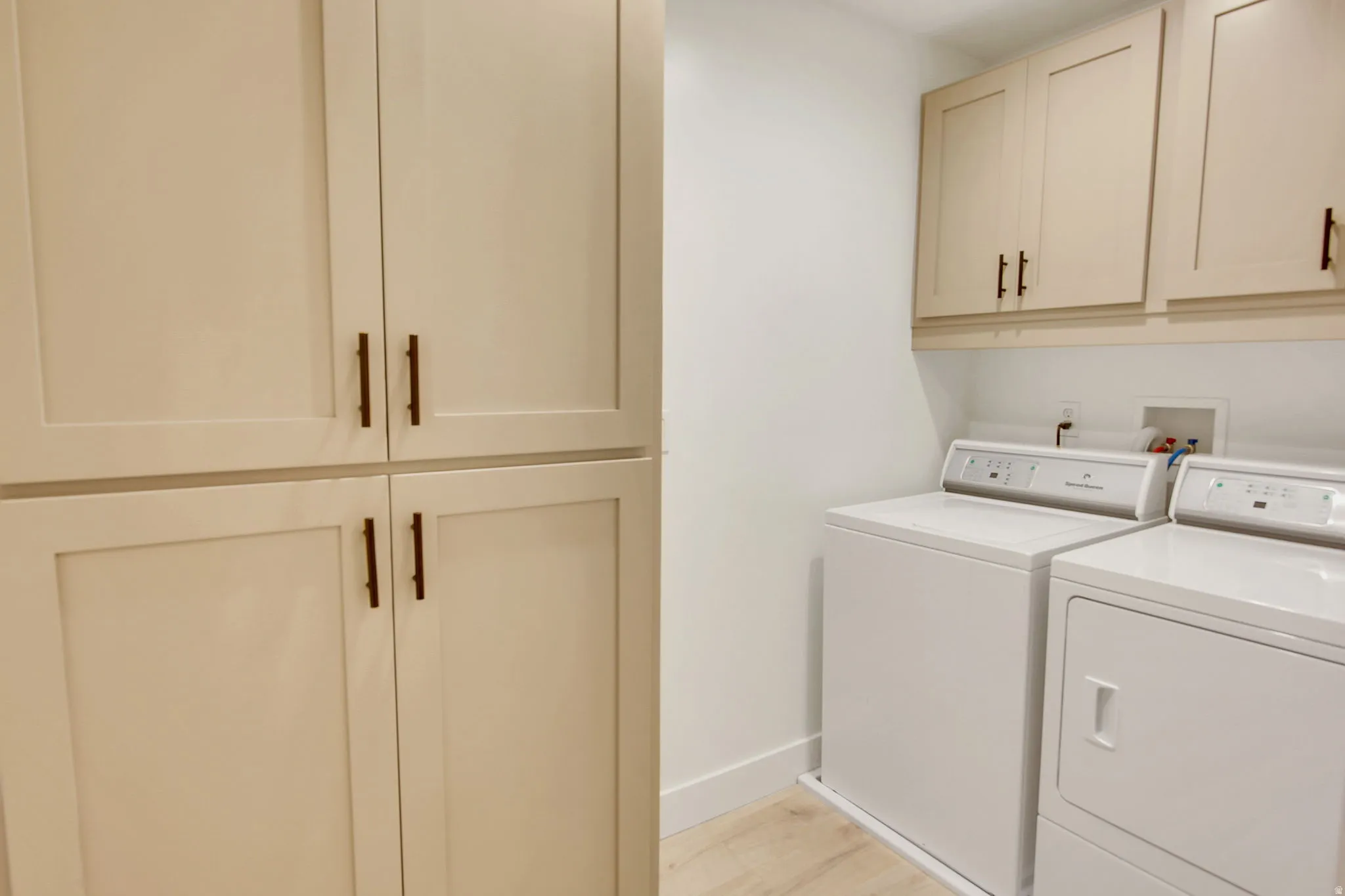 Laundry area featuring light-toned cabinetry with dark hardware, a pair of white appliances, and light wood-finish flooring