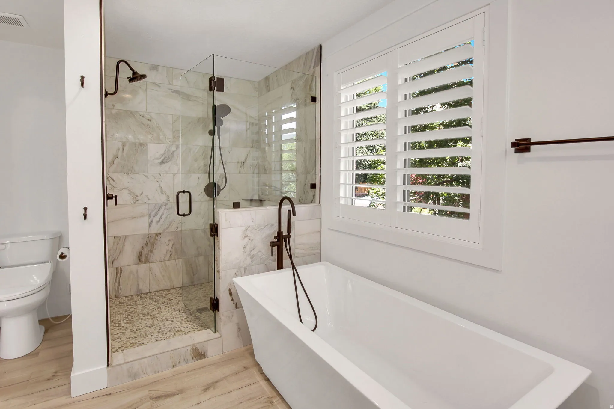 Contemporary bathroom featuring a freestanding soaking tub, a frameless glass shower enclosure with stone tile walls and a pebble-finish floor, and a window with plantation shutters