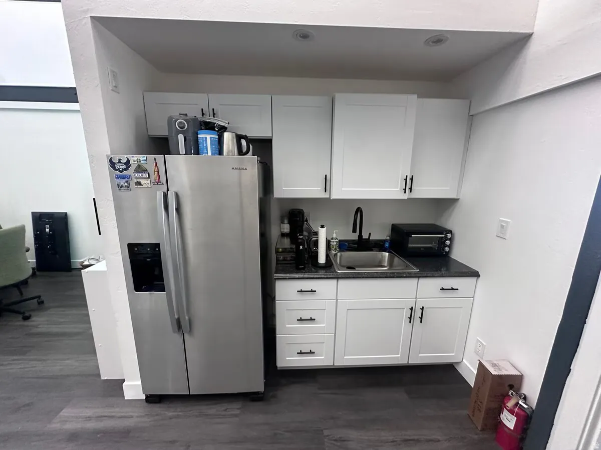 Compact kitchen area featuring white shaker cabinetry, dark granite-style countertops, an undermount sink with a matte black faucet, and stainless steel appliances including a side-by-side refrigerator