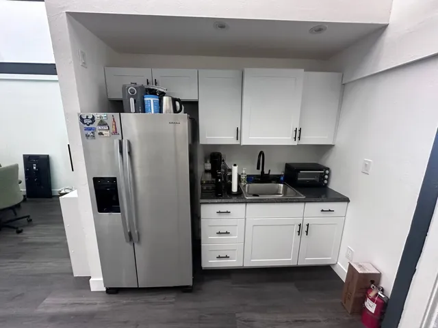 Compact kitchen area featuring white shaker cabinetry, dark granite-style countertops, an undermount sink with a matte black faucet, and stainless steel appliances including a side-by-side refrigerator
