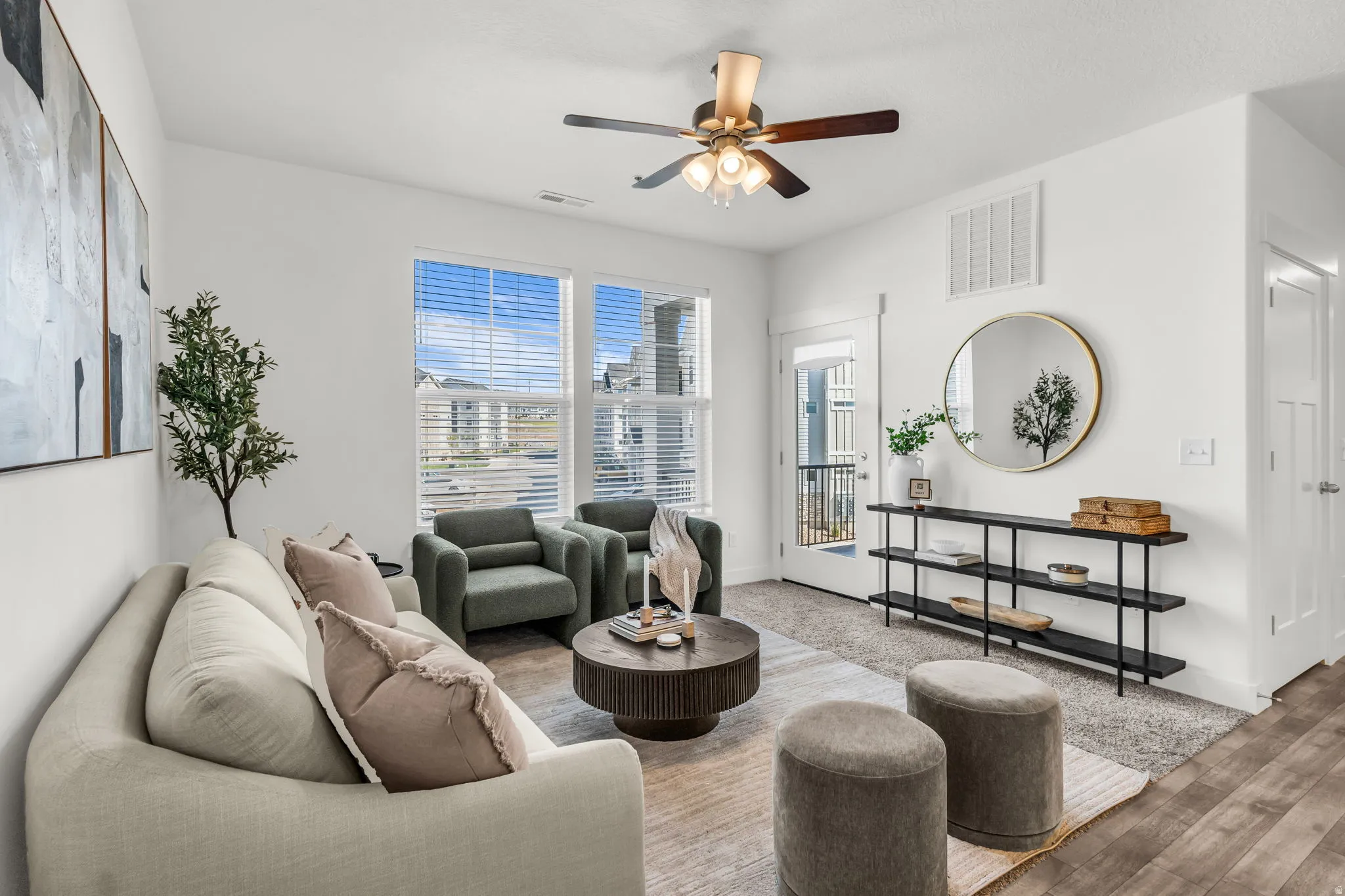 Bright living area featuring large windows with blinds, a ceiling fan with light fixture, wood-finish flooring extending from the entryway, a white panel door, and a glass-paneled exterior door