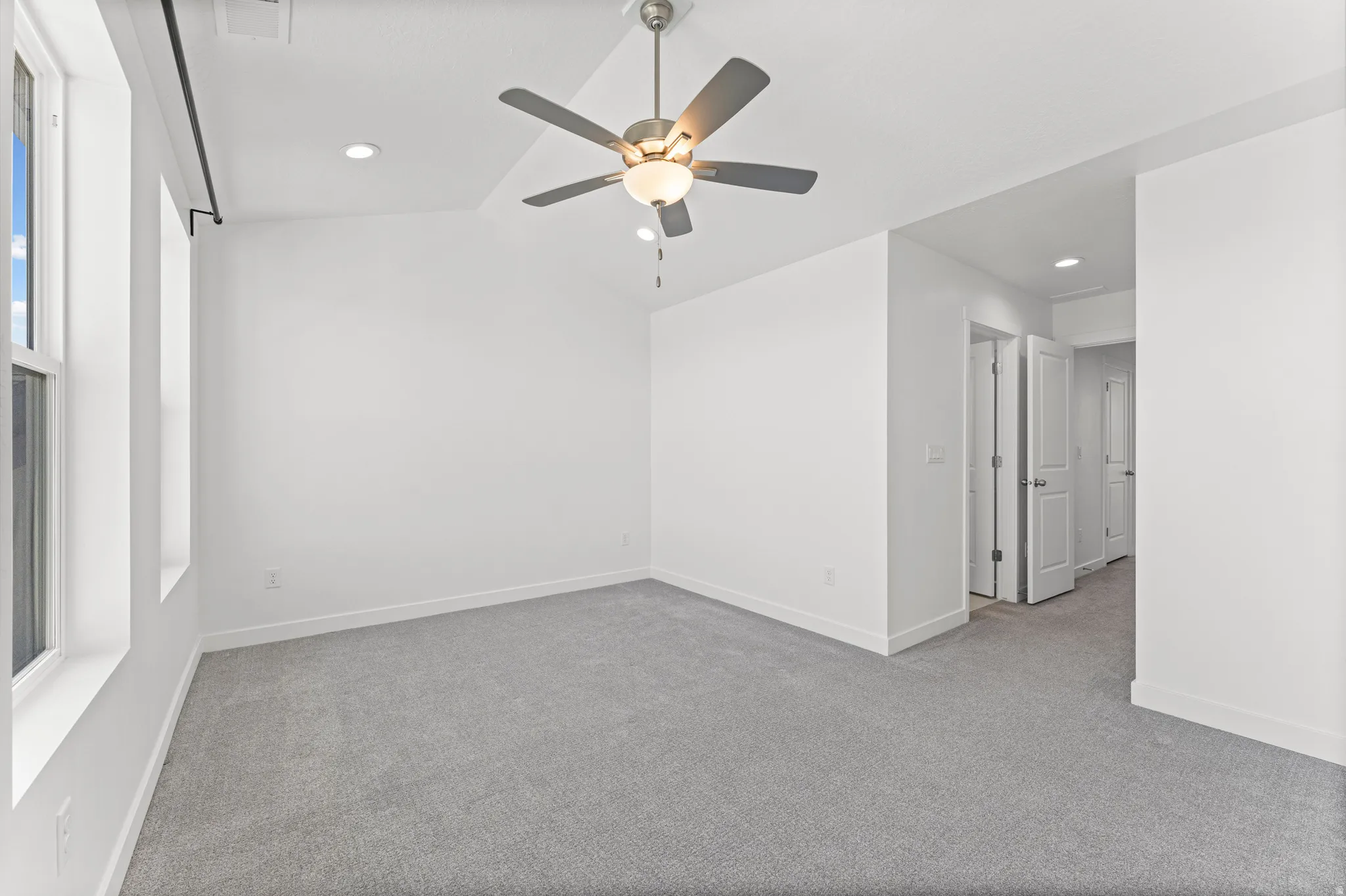 Spacious room featuring light gray carpeting, crisp white walls, multiple windows, a ceiling fan with integrated lighting, and recessed ceiling lights