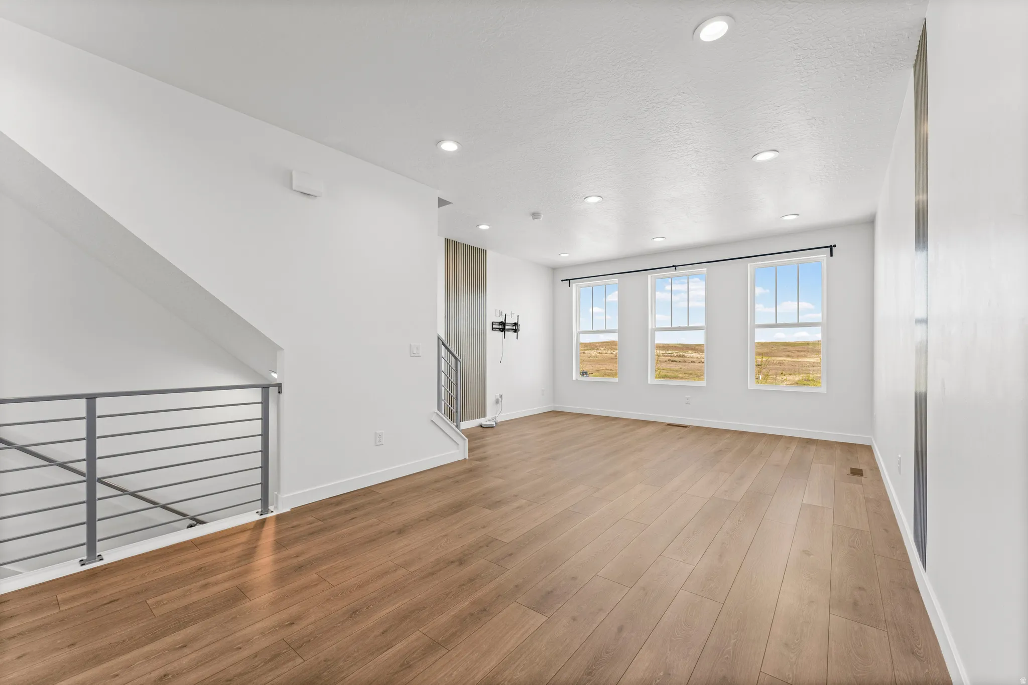 Spacious room featuring wood-finish flooring, a contemporary horizontal bar railing, three large windows, recessed ceiling lighting, and a white interior paint scheme