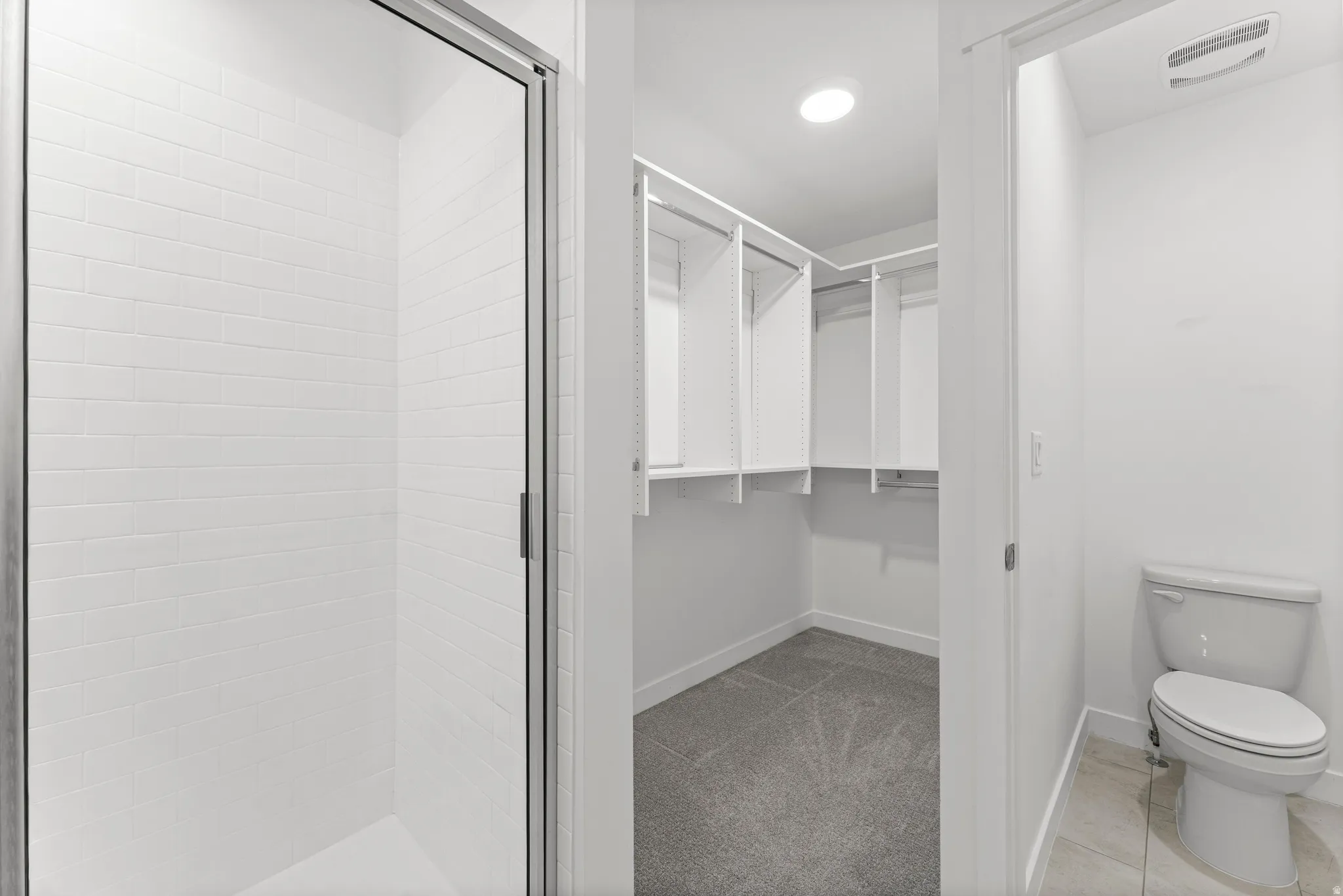 Walk-in shower with clear glass enclosure and white subway tile surround, alongside a spacious walk-in closet with built-in white shelving and gray carpet flooring