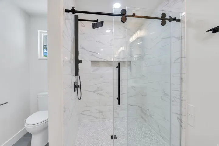 Frameless glass shower enclosure with matte black hardware, white subway tile walls with subtle veining, and white hexagonal mosaic floor tiles