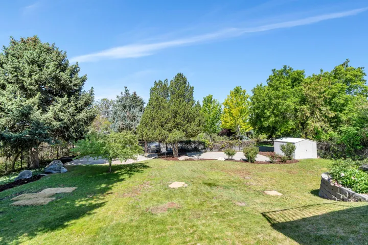 Expansive backyard featuring a manicured lawn, mature trees, landscaped beds, a stone-faced retaining wall, and a white storage shed