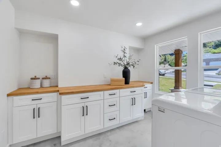 Bright utility space featuring dual windows, recessed lighting, wood-finish countertops, white cabinetry with black hardware, and an appliance bay