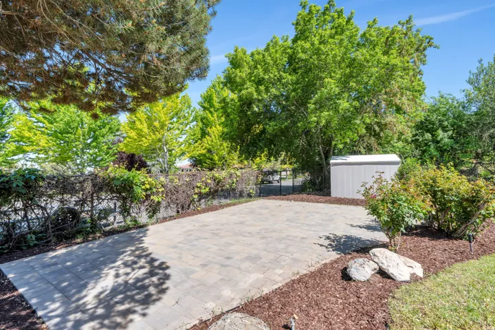 Paved patio area featuring interlocking pavers, surrounded by mature trees, landscaping with wood chips, and a storage shed