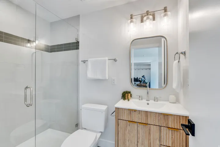Contemporary bathroom featuring a clear glass shower enclosure, a wood-finish vanity with gold-tone hardware, a rectangular vanity mirror, and modern three-light sconce