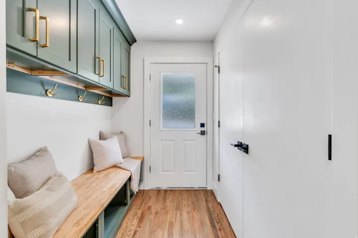 Entryway mudroom featuring wood-finish flooring, built-in bench seating, overhead cabinetry, wall-mounted hooks, and a white door with frosted glass