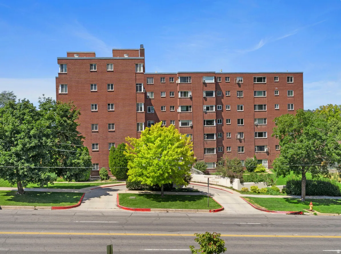 Multi-story brick building featuring numerous windows, a visible rooftop section, and a manicured front lawn with mature trees