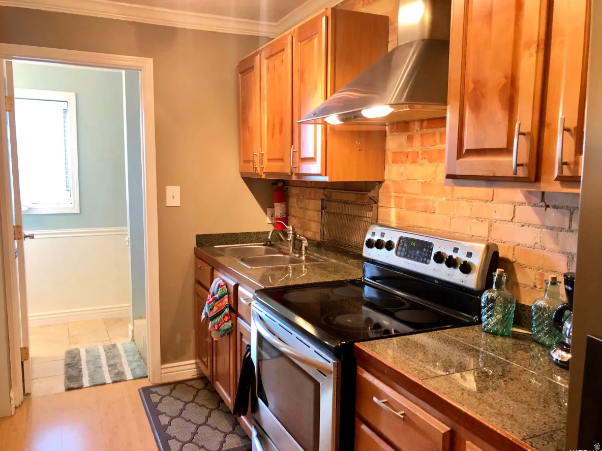 Kitchen featuring wood cabinetry, a stainless steel range hood, stone countertops, a brick-finish backsplash, and wood-finish flooring
