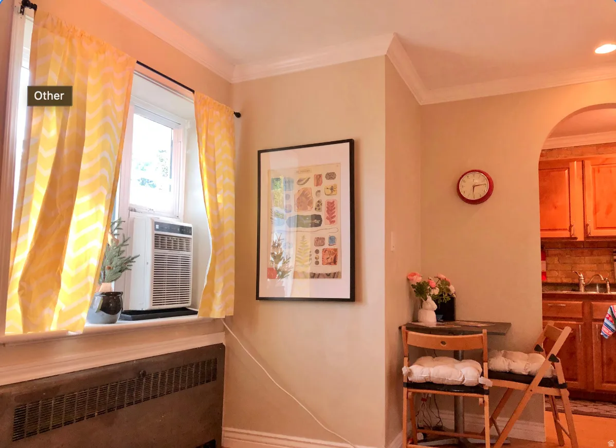 Crown molding and neutral wall paint define this living space, featuring a window with natural light, a baseboard radiator, and an arched doorway leading into a kitchen with wood cabinetry and a tile backsplash