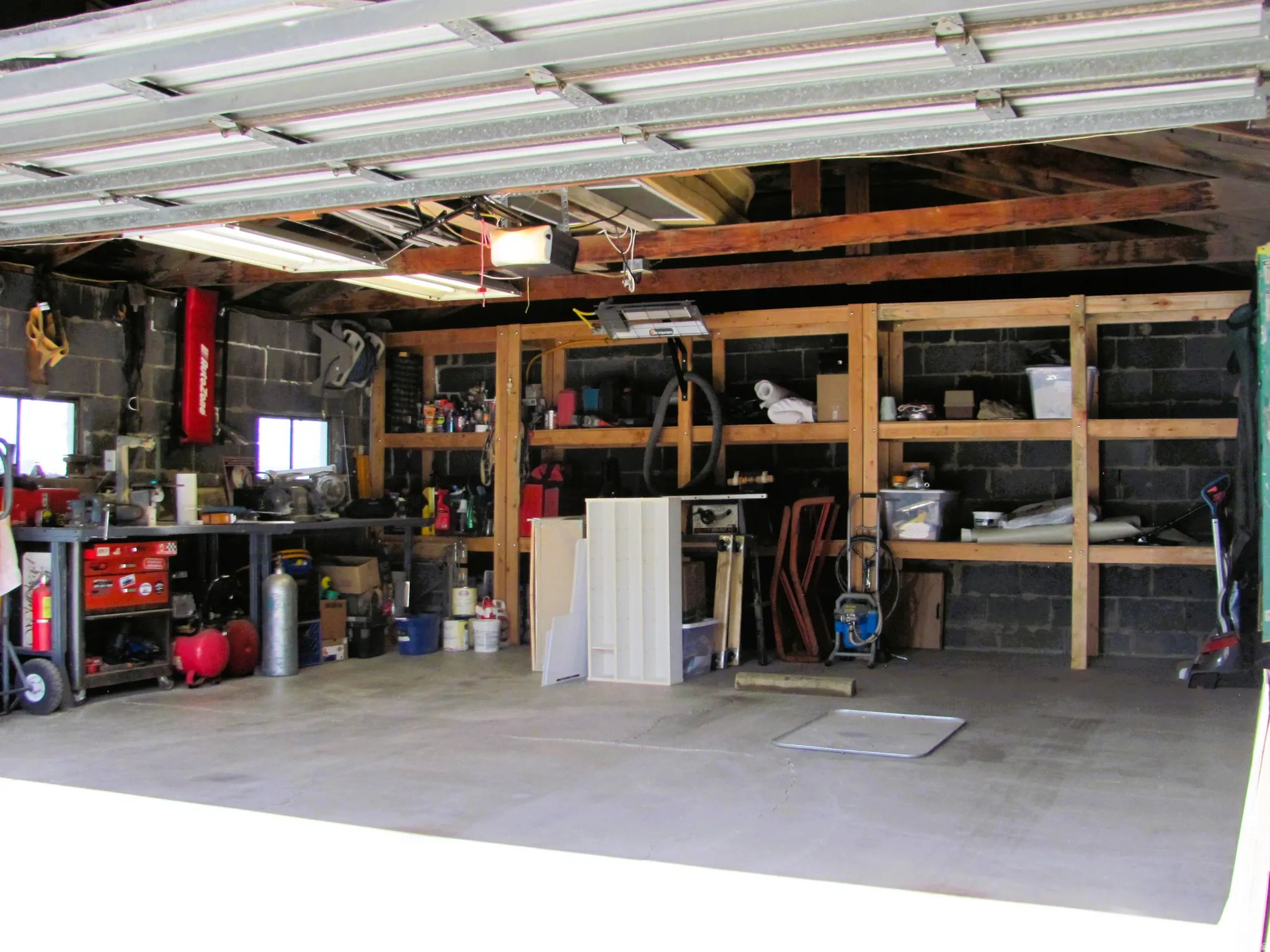 Spacious garage featuring a concrete floor, exposed ceiling beams, built-in shelving, two windows, and fluorescent lighting