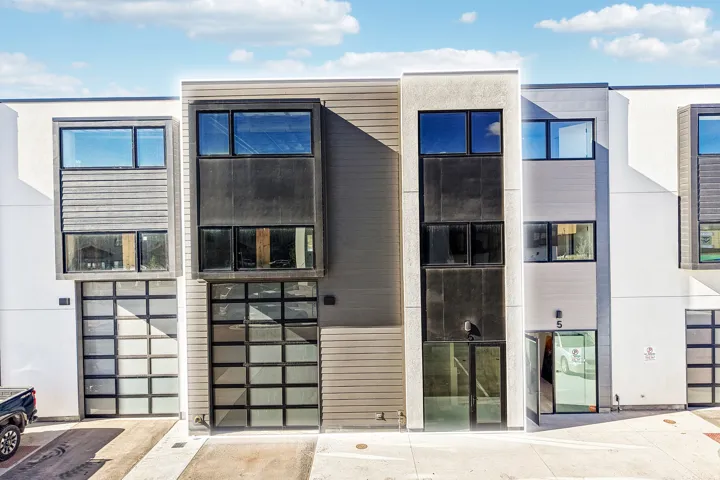 Modern townhome facade featuring a prominent glass-paneled garage door, large rectangular windows, and a multi-textured exterior combining light-colored stucco and dark horizontal siding