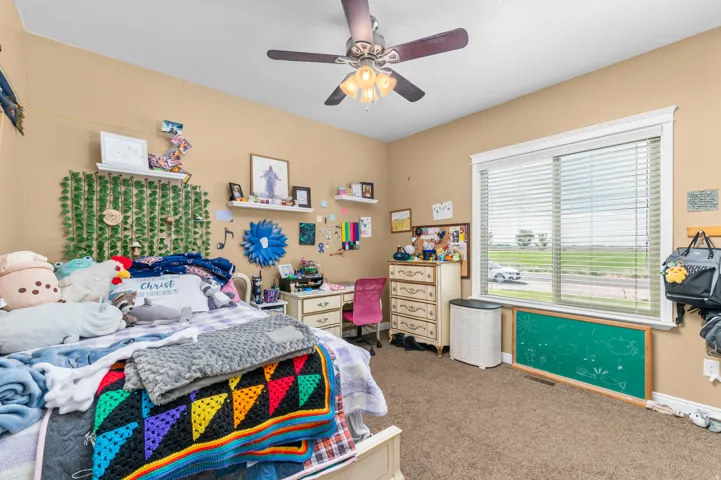 Spacious bedroom featuring light brown carpeting and walls, a ceiling fan with integrated lighting, and a large window with white blinds and trim