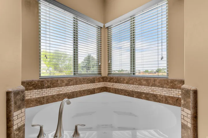 Corner soaking tub featuring brushed nickel fixtures, a tiled surround with a mosaic accent band, and two large windows with blinds