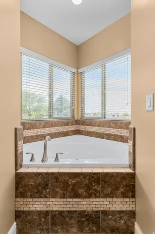 Corner soaking tub featuring brushed nickel fixtures, a dark brown tiled surround with a light mosaic accent band, and perimeter windows with horizontal blinds