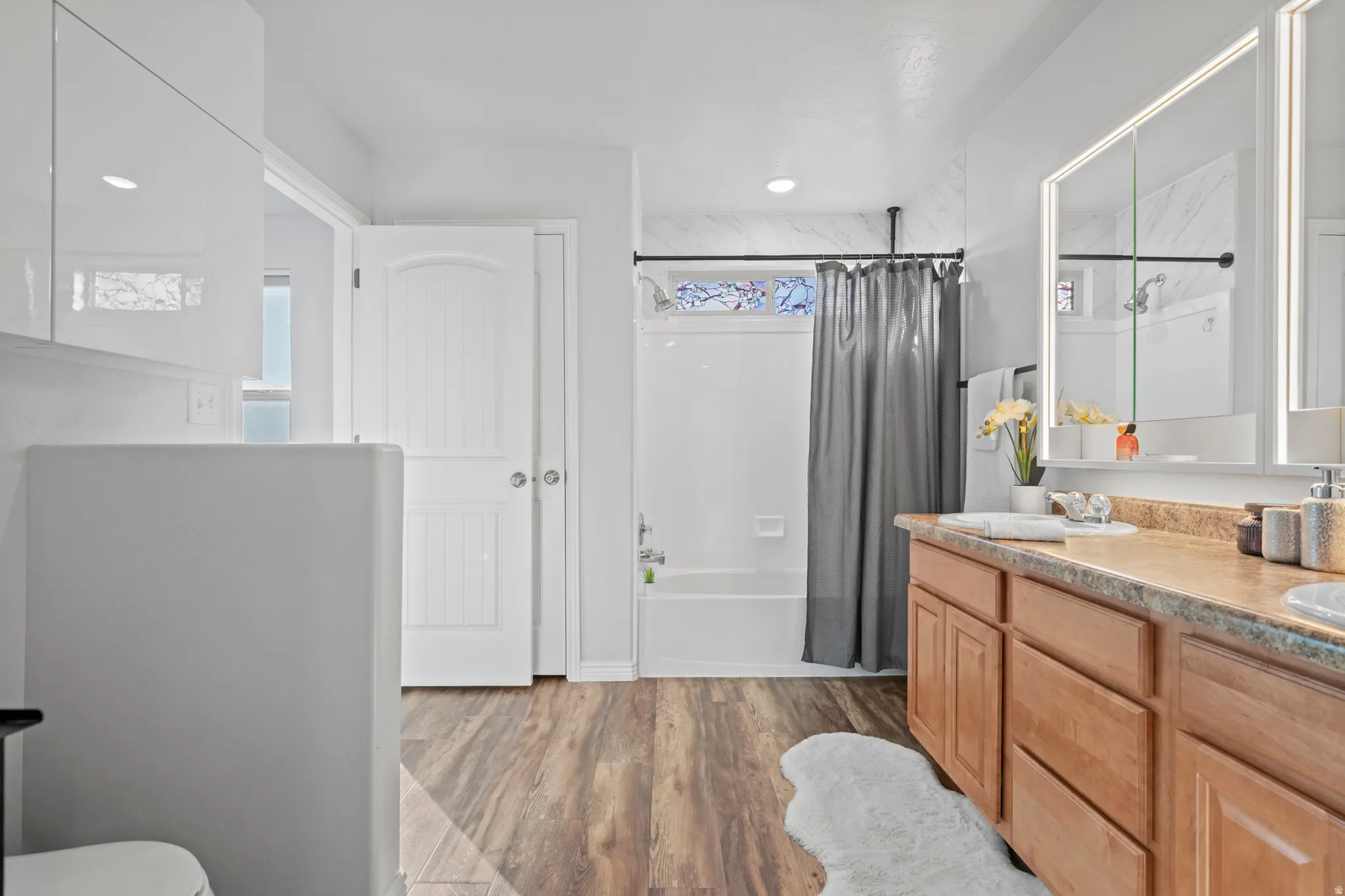 Bathroom featuring a dual vanity, wood-finish flooring, a white bathtub with shower, and modern illuminated medicine cabinets