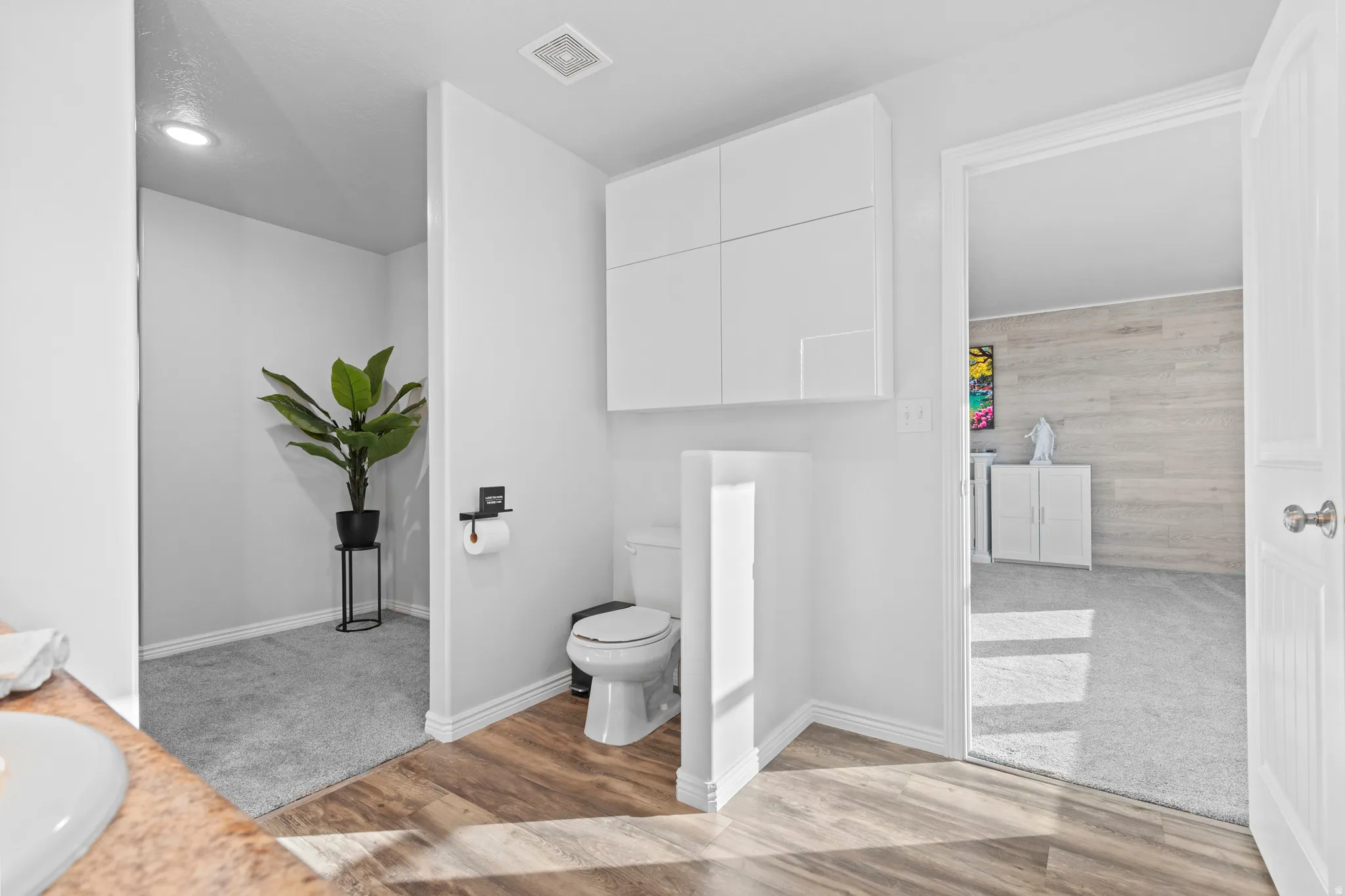 Bathroom featuring wood-finish flooring, a white toilet with an enclosed privacy wall, and upper white cabinetry